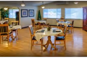 A dining room with several round tables covered with white tablecloths, each set with plates, napkins, and utensils. Wooden chairs surround the tables. The room has large windows letting in natural light, framed pictures on the walls, and a wooden floor.