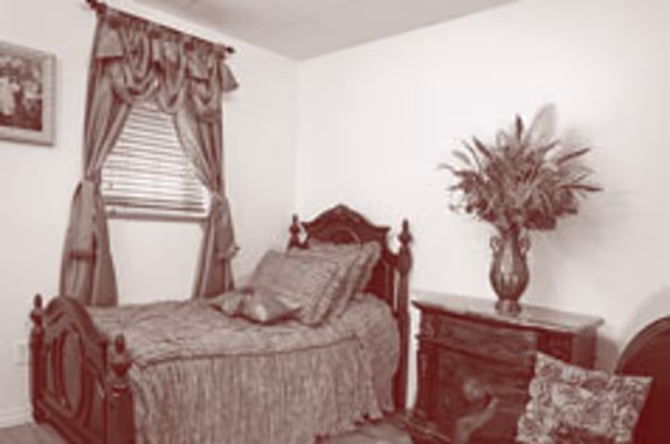 A bedroom with a single wooden bed covered with a textured bedspread and pillows. Next to the bed is a wooden dresser with a large vase holding a floral arrangement. A window with draped curtains is on the wall behind the bed, and a framed picture hangs on the adjacent wall.