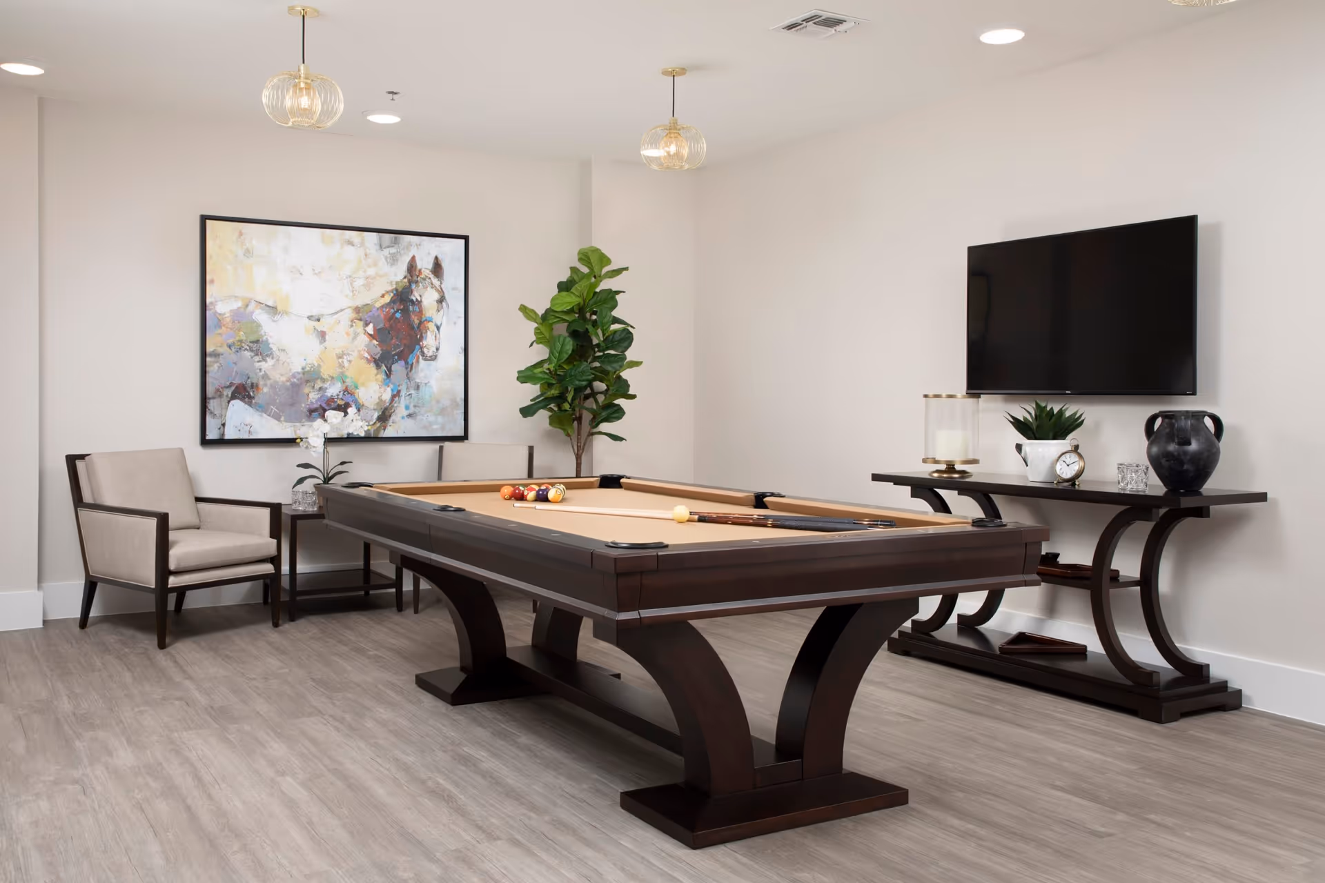 A bright interior recreation room with a pool table, chairs, wall art, and a wall-mounted TV on a console.