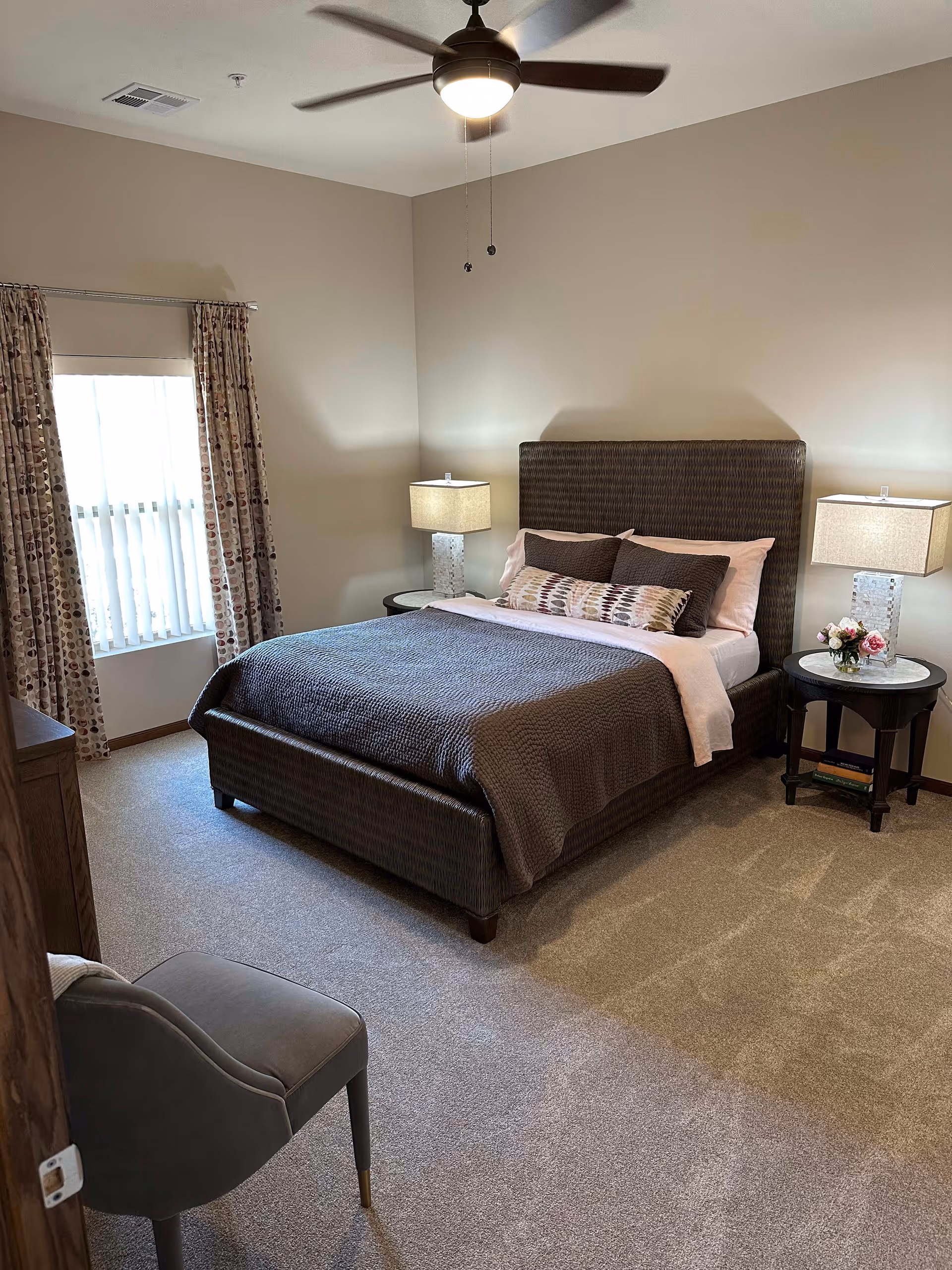 A neatly arranged bedroom with a large bed featuring a dark textured headboard and bedding. There are two bedside tables with matching lamps and a small flower arrangement on one table. A window with patterned curtains allows natural light into the room. A ceiling fan with a light is mounted above, and a gray upholstered chair is positioned near the door.