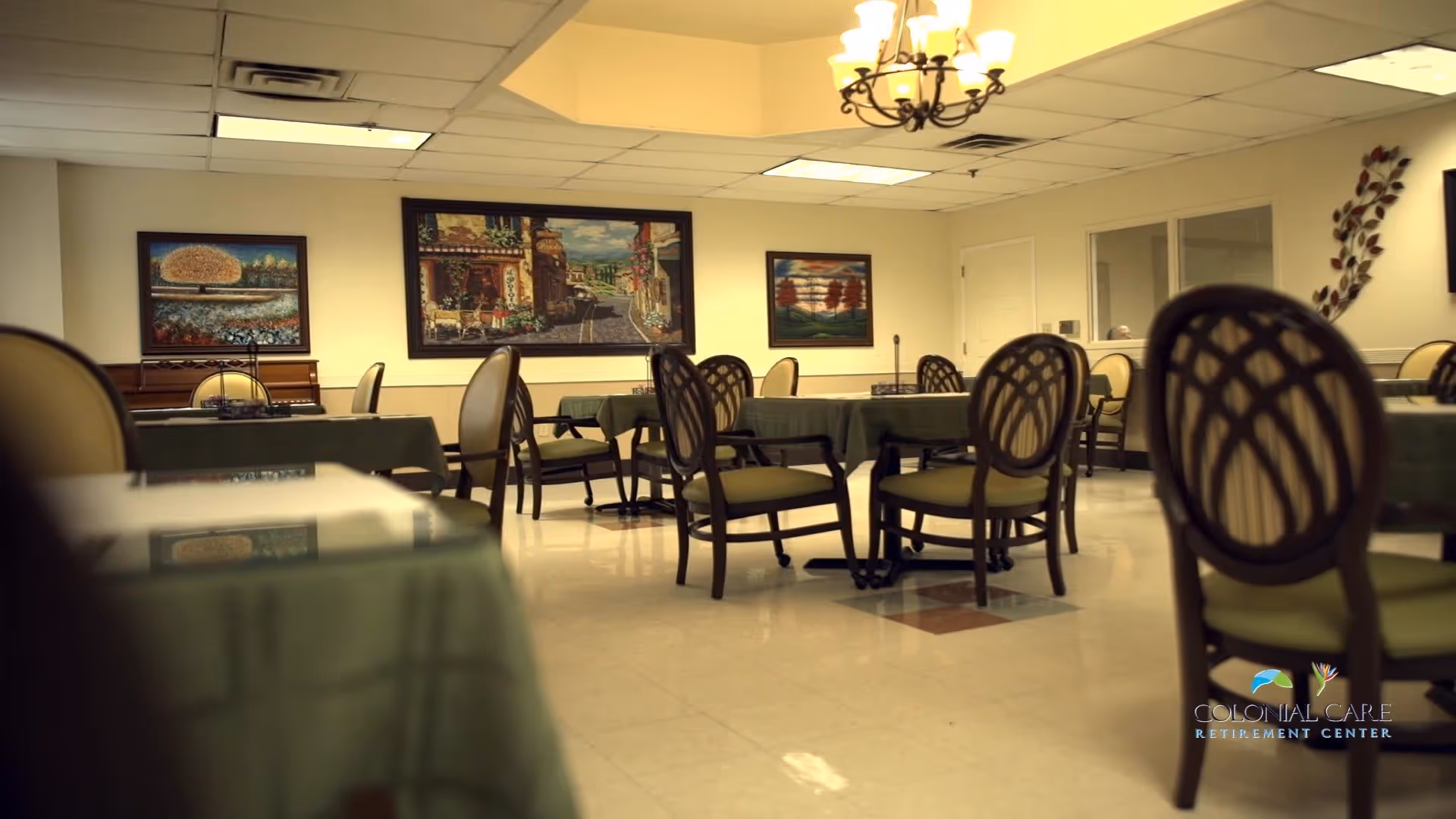 A dining room with multiple tables covered with green tablecloths and surrounded by wooden chairs with cushioned seats. The room has a tiled floor, a chandelier hanging from the ceiling, and framed paintings on the walls. There is a windowed partition on one side and a decorative wall piece.