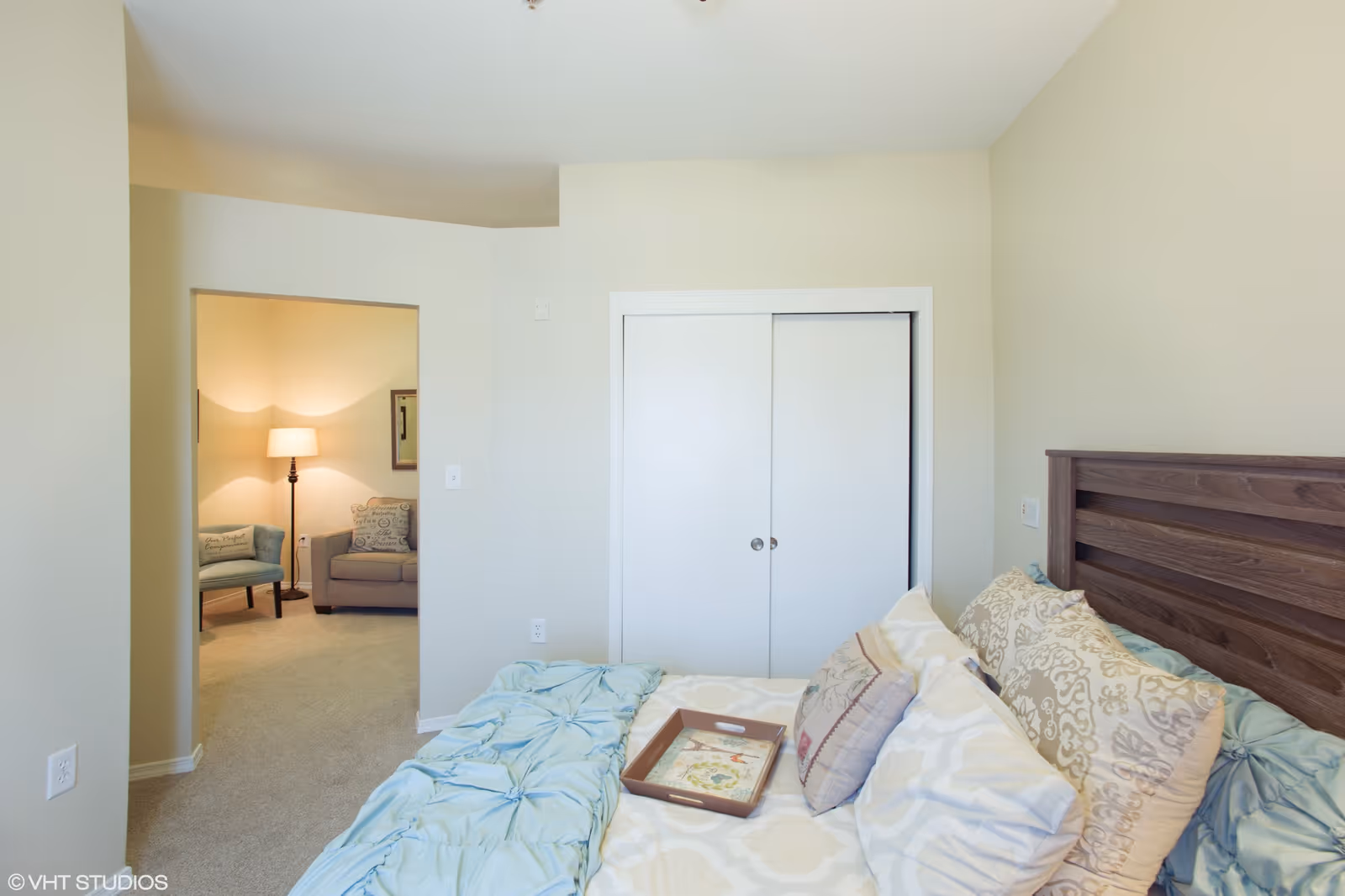 A bedroom with a wooden headboard, a bed covered with a light blue and beige comforter and multiple pillows. A wooden tray is placed on the bed. The room has light-colored walls and carpeted floor. There is a white double-door closet and an open doorway leading to a sitting area with a floor lamp, a gray sofa, and a blue armchair.