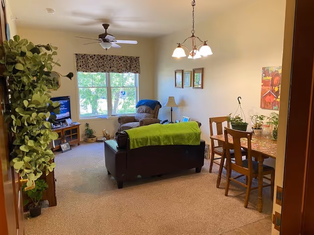 A cozy senior living room with a brown leather sofa covered partially with a green blanket, a recliner chair near a window with floral curtains, a wooden TV stand with a television, and a dining table with four chairs and potted plants on top. The room is carpeted and has a ceiling fan and a hanging light fixture.