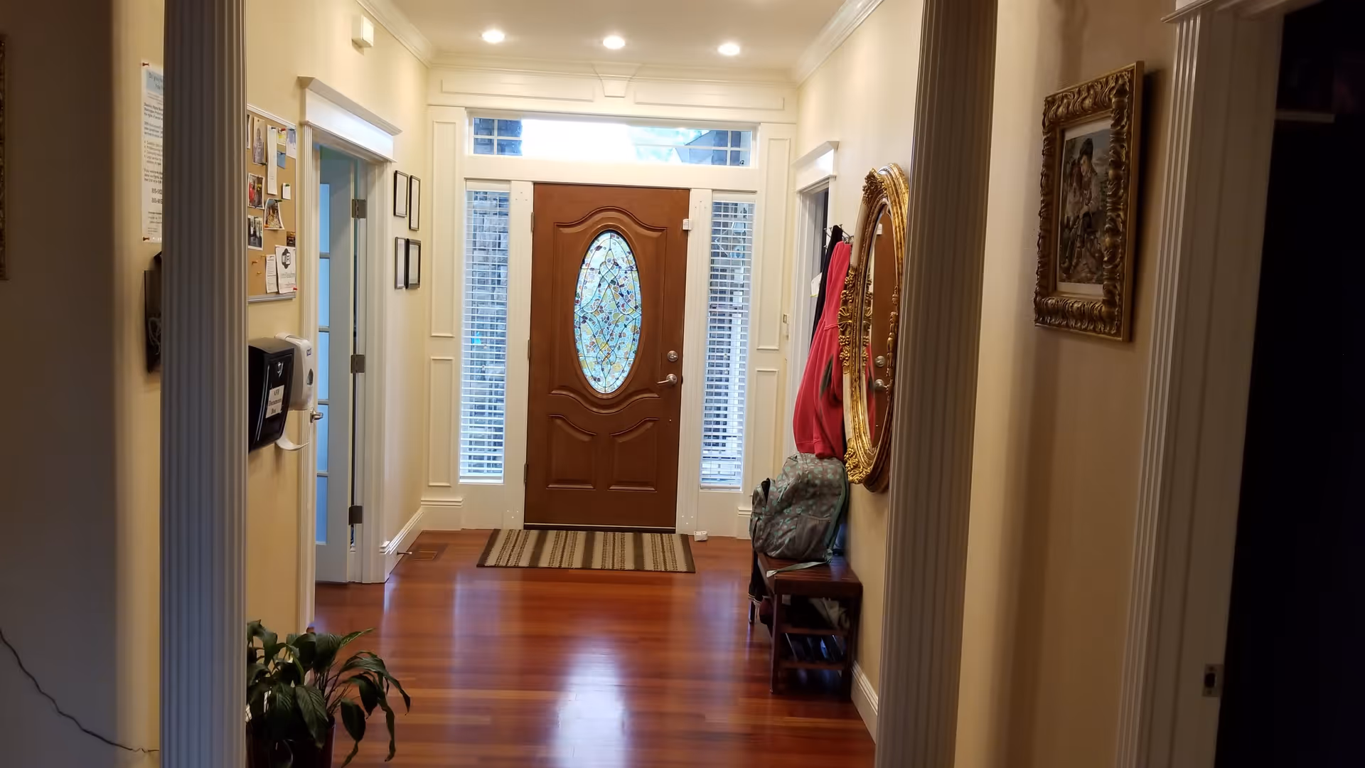 Interior view of a hallway with polished wooden floors leading to a wooden front door with an oval stained glass window. The hallway has cream-colored walls, a large ornate gold-framed mirror on the right wall, a small bench with bags and coats hanging above it, and a bulletin board on the left wall near an open doorway.