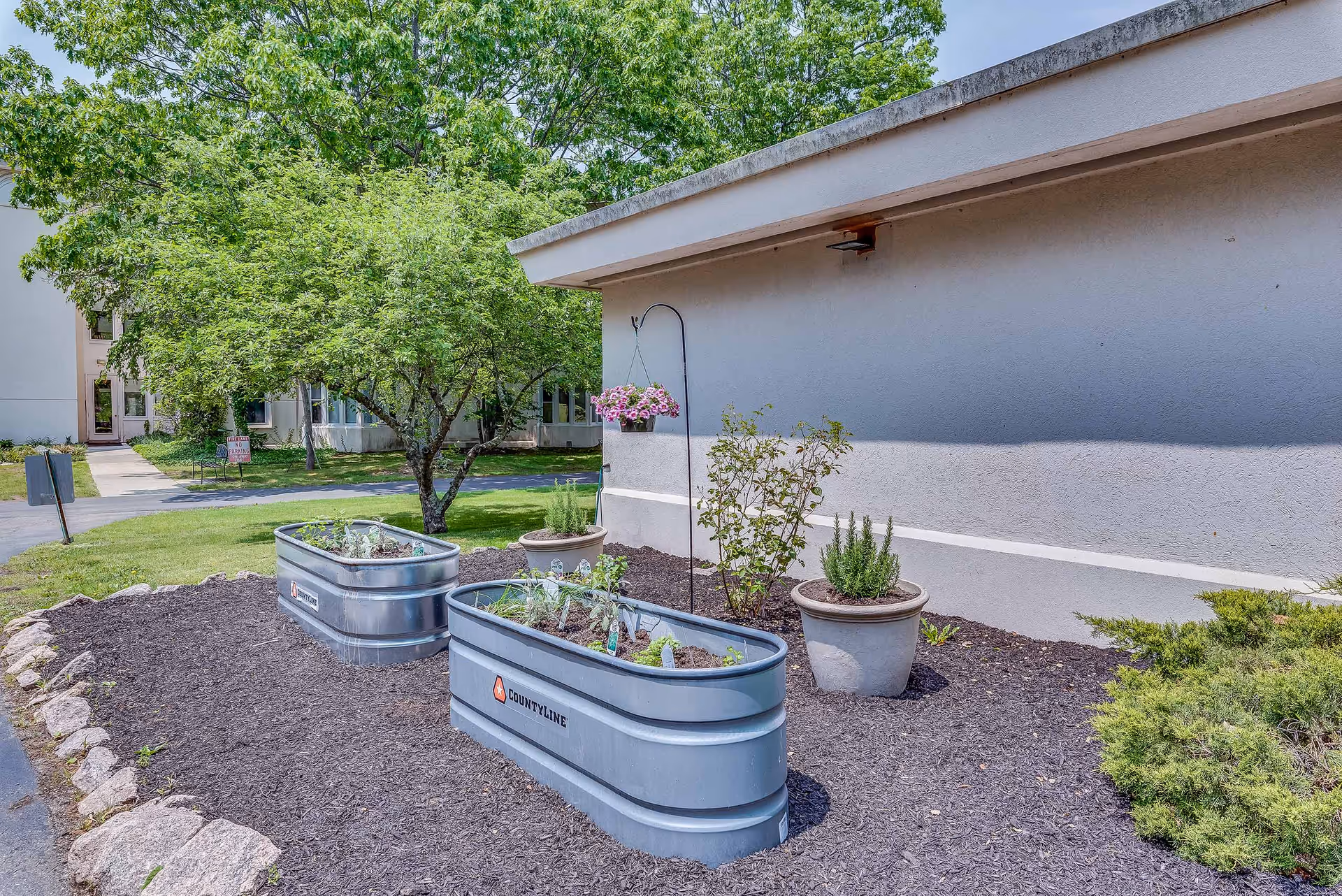 Outdoor garden area at Guilford Retirement Residence featuring two raised metal garden beds with plants, potted plants, a hanging flower basket, a tree, and a building wall in the background.