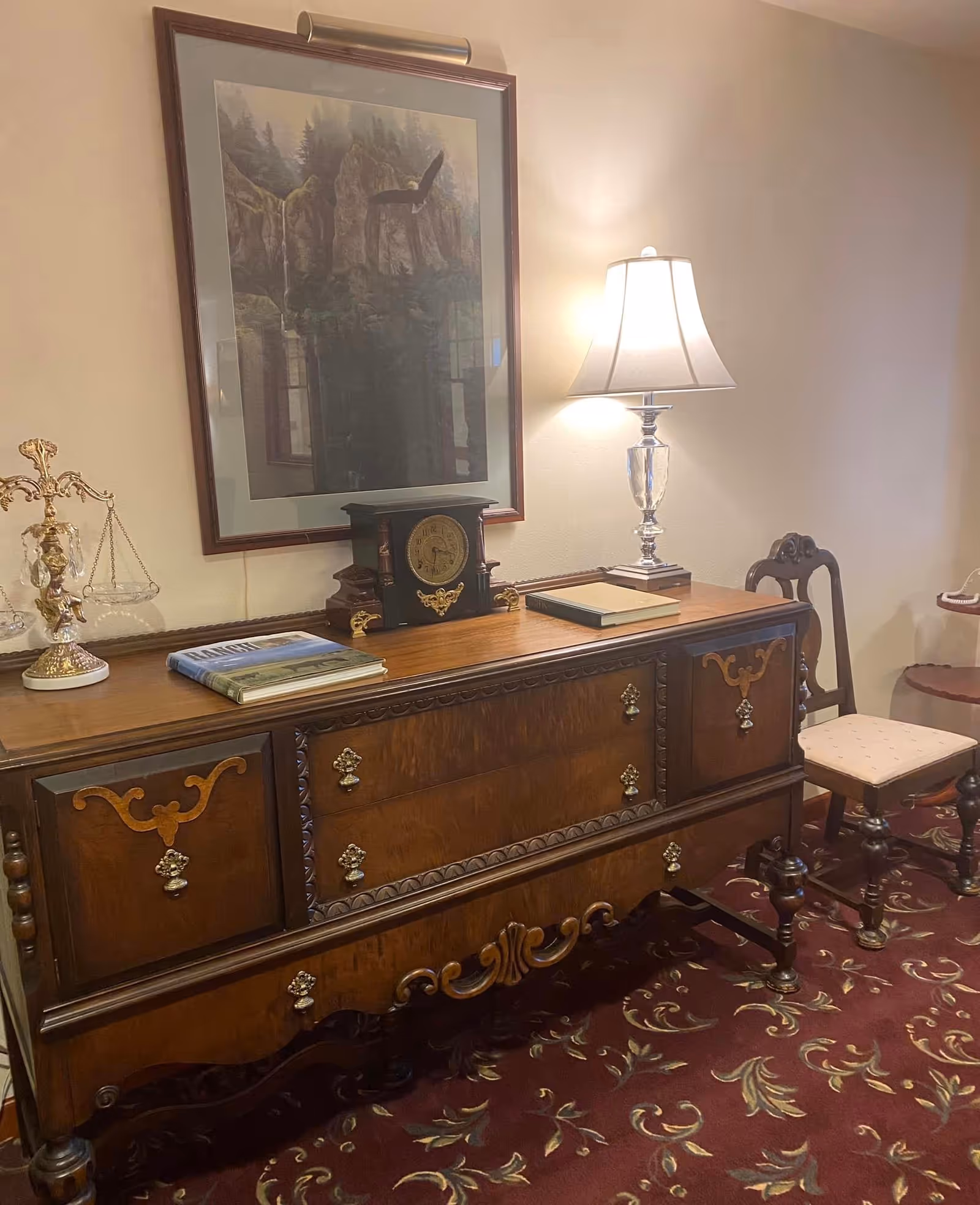 An antique wooden sideboard with ornate carvings and brass handles, topped with a vintage clock, a decorative scale, two books, and a table lamp with a white shade. Above the sideboard hangs a framed painting of a waterfall and forest scene. To the right, there is a wooden chair with a cushioned seat and a small round table. The floor is covered with a patterned carpet.