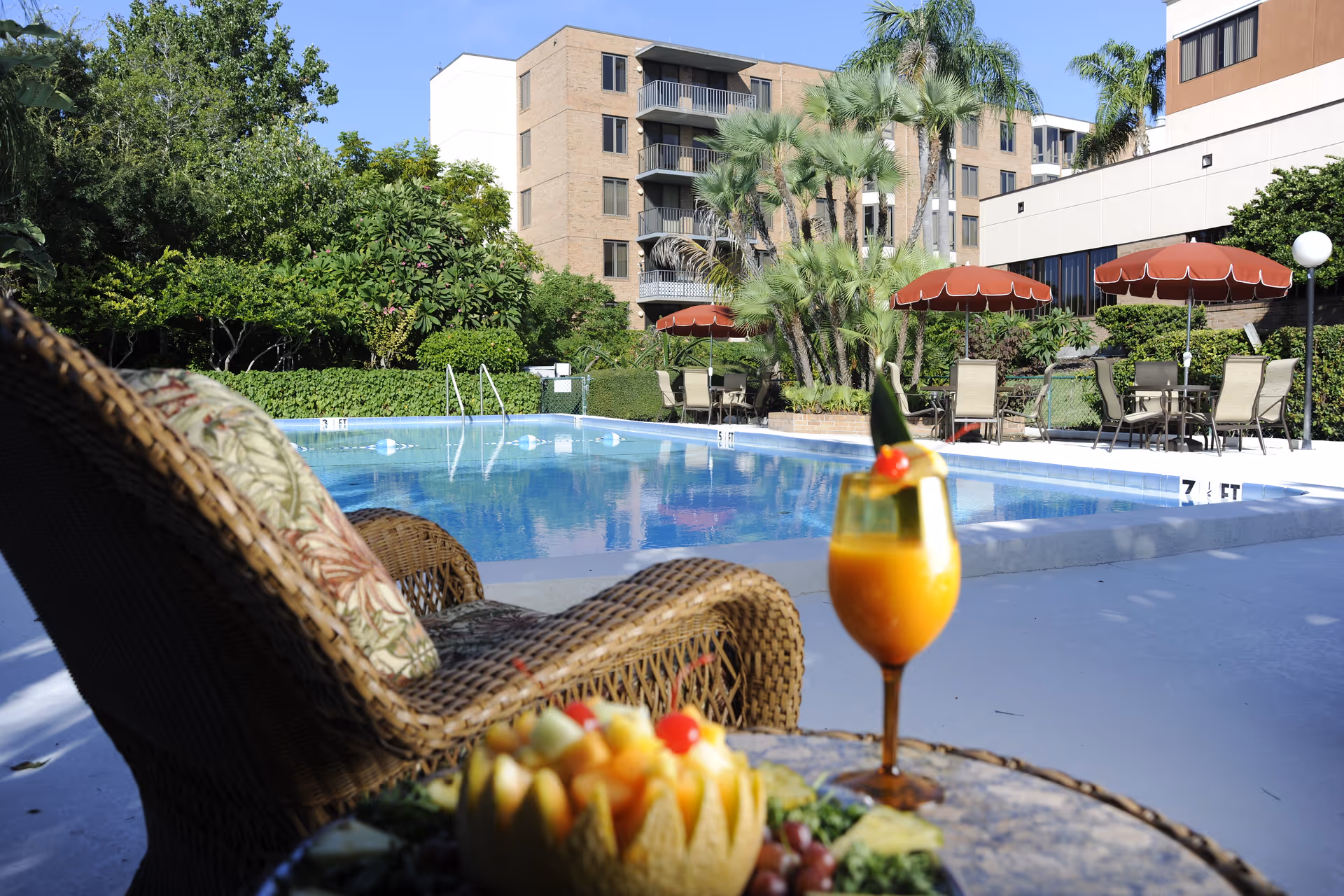 Outdoor swimming pool area with lounge chairs and tables shaded by red umbrellas. There are palm trees and other greenery surrounding the pool. In the foreground, there is a wicker chair with a floral cushion and a table holding a tropical drink and a fruit platter. Apartment buildings are visible in the background under a clear blue sky.