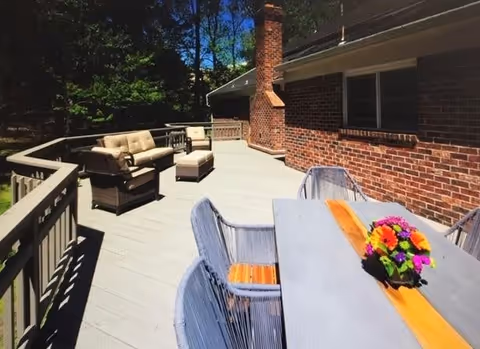 Outdoor deck area with a wooden railing, featuring a gray table with a colorful flower arrangement and four gray chairs. In the background, there is a cushioned seating area with a sofa, two chairs, and an ottoman. The deck is attached to a brick building with a chimney, surrounded by trees and greenery under a clear blue sky.