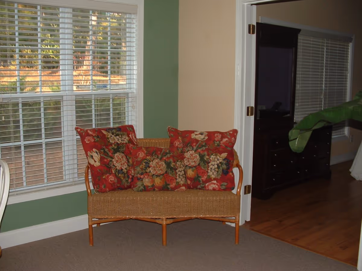 A wicker bench with red floral cushions sits by a large window in a home interior with a doorway leading to a bedroom.
