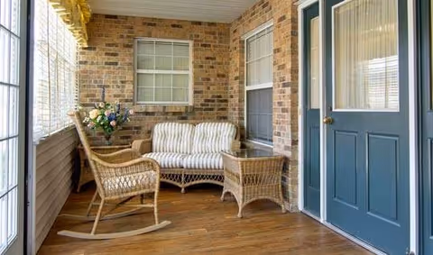 A cozy enclosed porch with wooden flooring and brick walls, featuring a wicker rocking chair, a wicker loveseat with striped cushions, a wicker side table, a bouquet of flowers in a vase, and a blue door with glass panels.
