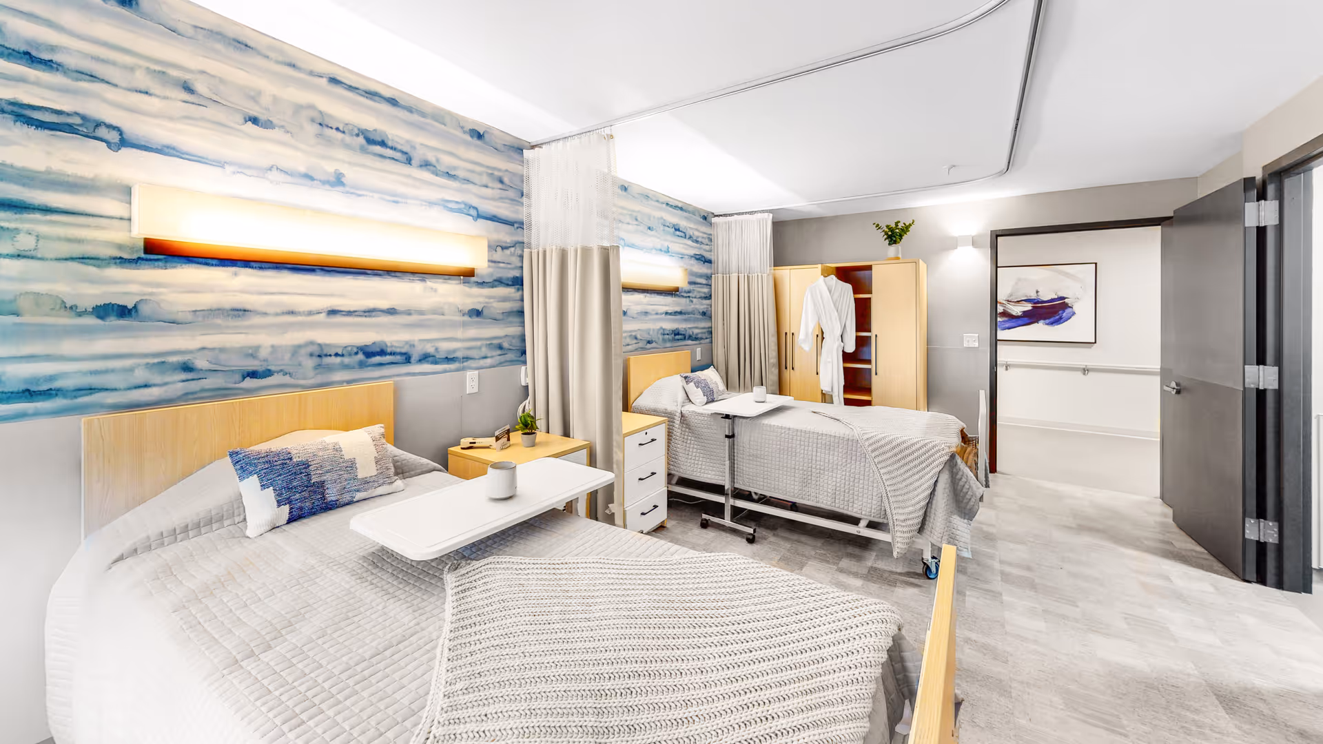A bright and clean rehabilitation room with two hospital-style beds, each with a small overbed table and a pillow. The room features a blue and white abstract wall design, light wood furniture including a nightstand and a wardrobe with a white robe hanging inside. The floor is light-colored, and there is a door open to a hallway with a framed artwork on the wall.