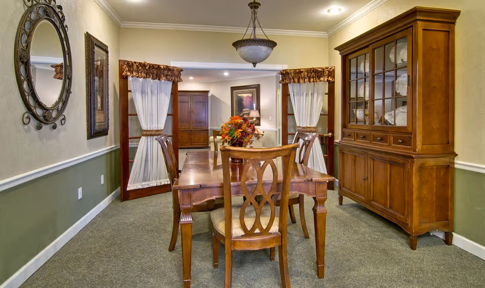 A traditional dining room with a wooden dining table and four chairs. The table has a floral centerpiece. There is a wooden china cabinet on the right side filled with dishes. The room has beige walls with a green lower half and white trim. Two French doors with sheer white curtains and brown valances lead to another room with a framed picture and lamp visible. A decorative round mirror and framed artwork hang on the left wall.
