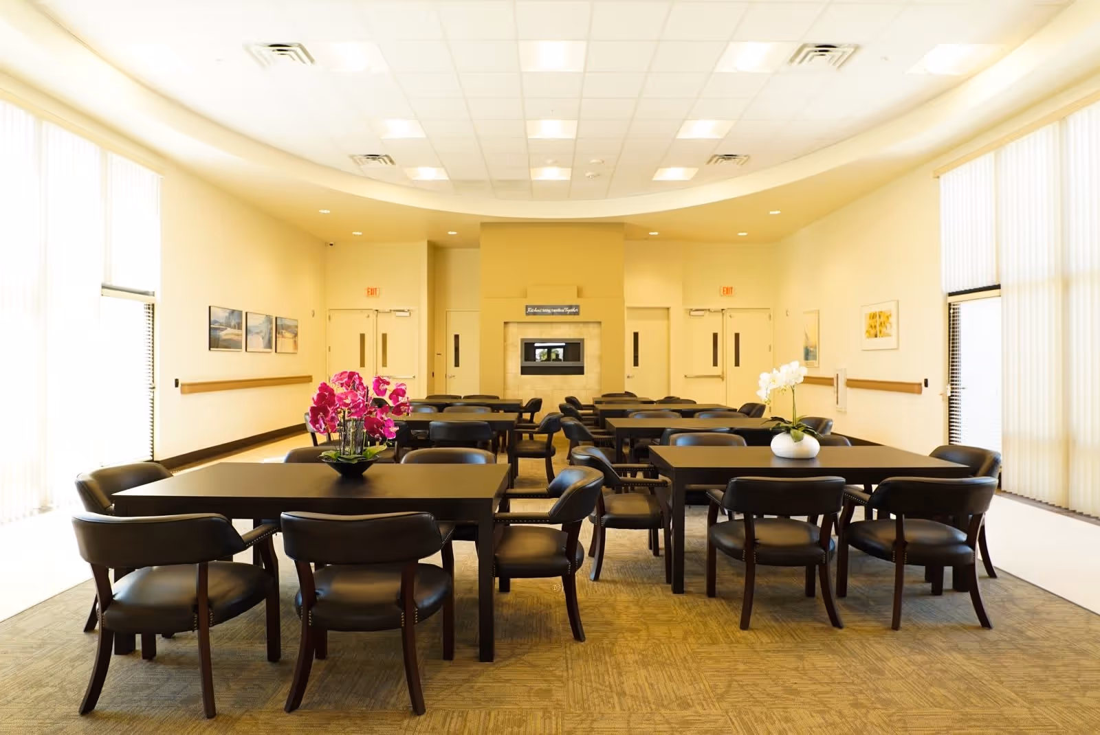 A bright dining room with multiple dark wood tables and black chairs arranged neatly. Each table has a floral centerpiece with pink or white flowers. The room has large windows with vertical blinds allowing natural light to fill the space. The walls are light-colored with framed artwork, and there is a fireplace or built-in feature at the far end of the room.