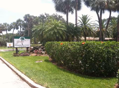 Sunlit entrance area with palm trees, trimmed hedges, and a facility sign.