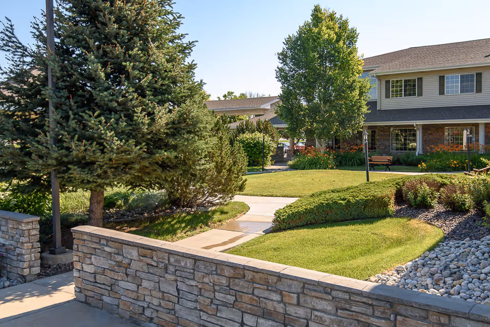 Outdoor view of a senior living facility with a stone wall in the foreground, a paved walkway, green grass, bushes, trees, and a two-story building with large windows in the background under a clear blue sky.