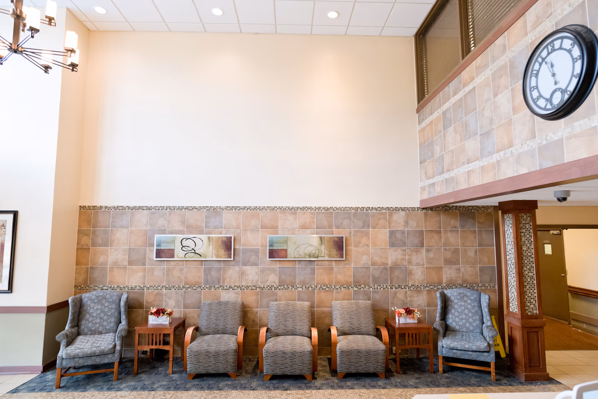 A seating area in a healthcare facility with five chairs arranged in a row against a tiled wall. Two patterned armchairs are on each end with three cushioned chairs with wooden armrests in the middle. There are two small wooden side tables with flower arrangements between the chairs. Above the chairs, two abstract paintings hang on the tiled wall. A large clock is mounted on the upper right wall, and a chandelier is partially visible on the upper left. The area is well-lit with a high ceiling and recessed lighting.