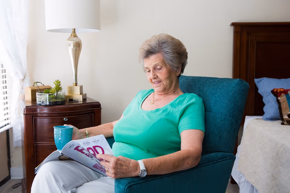 An elderly woman with short gray hair wearing a green shirt sits in a teal armchair in a bedroom. She is holding a magazine and a blue mug, with a wooden nightstand and lamp beside her and a bed with pillows in the background.