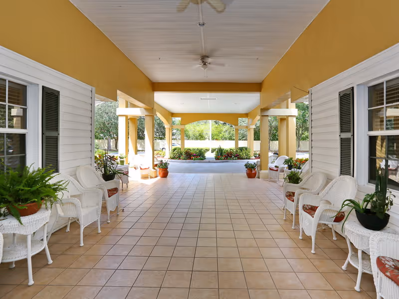 Covered outdoor walkway with tiled floor, white wicker chairs with cushions, and small tables holding potted plants. The walkway is flanked by white walls with windows and green shutters, leading to a garden area with more plants and trees visible in the background.