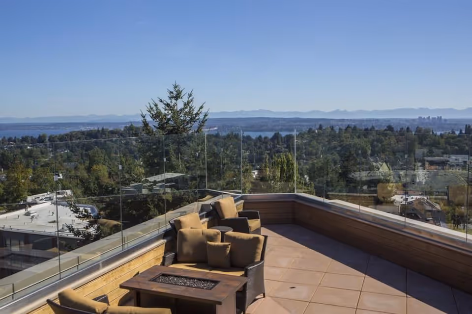 Outdoor patio area with cushioned chairs and a fire pit table, surrounded by glass railings overlooking a scenic view of trees, water, and distant mountains under a clear blue sky.