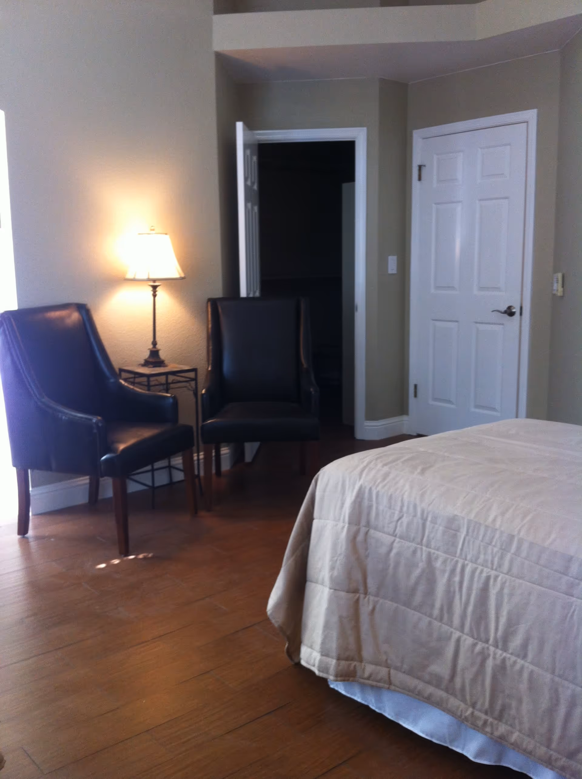 Bedroom corner with a beige-covered bed, two black leather chairs, a floor lamp, and a doorway with closed white doors.