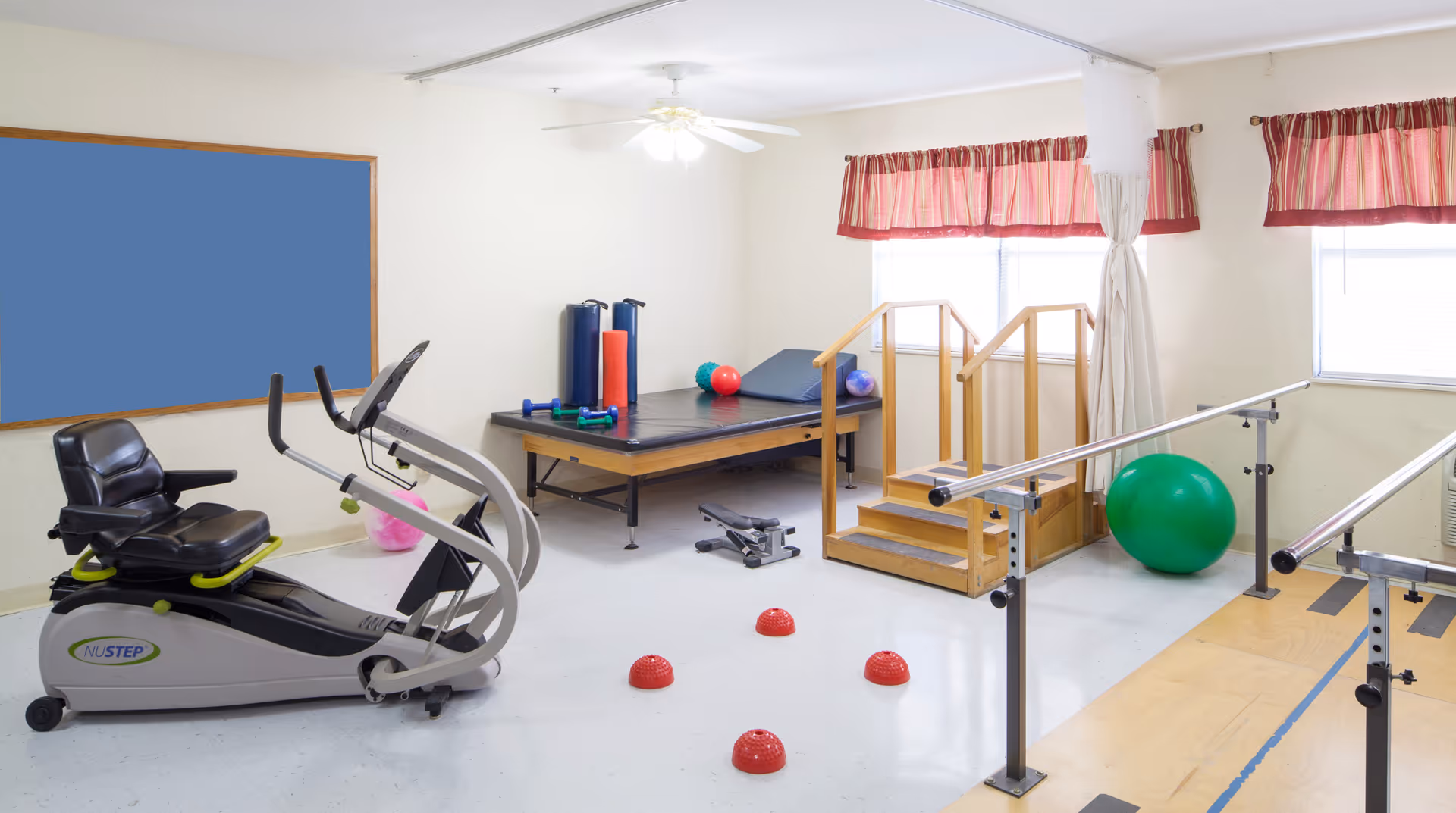 Therapy/exercise room with a recumbent exercise bike, parallel bars, a treatment table, therapy steps, and various exercise balls and balance equipment.