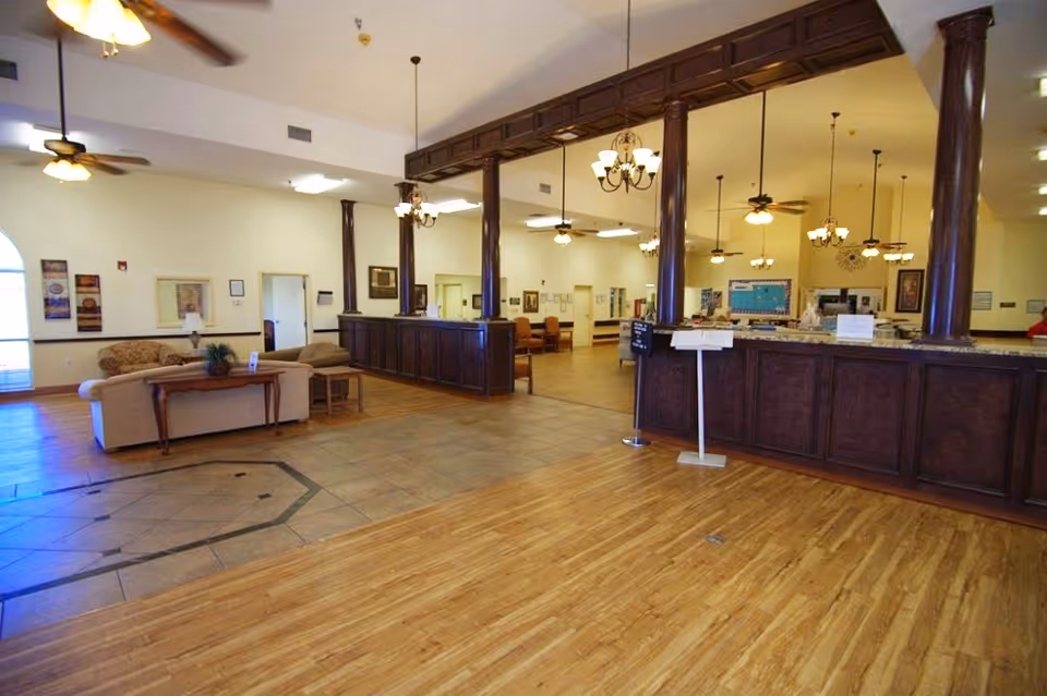 Spacious interior of a senior living facility with wooden flooring and tiled area near the entrance. There is a large dark wood reception desk with columns and chandeliers hanging from the ceiling. Comfortable seating with sofas and a table is visible on the left side, along with framed artwork on the walls. The area is well-lit with ceiling fans and lights.