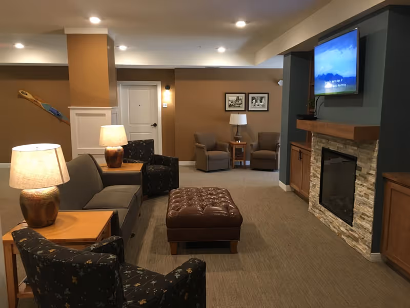 A cozy senior living room with brown walls and carpeted floor. The room features a stone fireplace with a mounted flat-screen TV above it, a brown leather tufted ottoman in the center, and several upholstered chairs and sofas arranged around wooden side tables with lamps. Two framed pictures hang on the far wall near a white door.