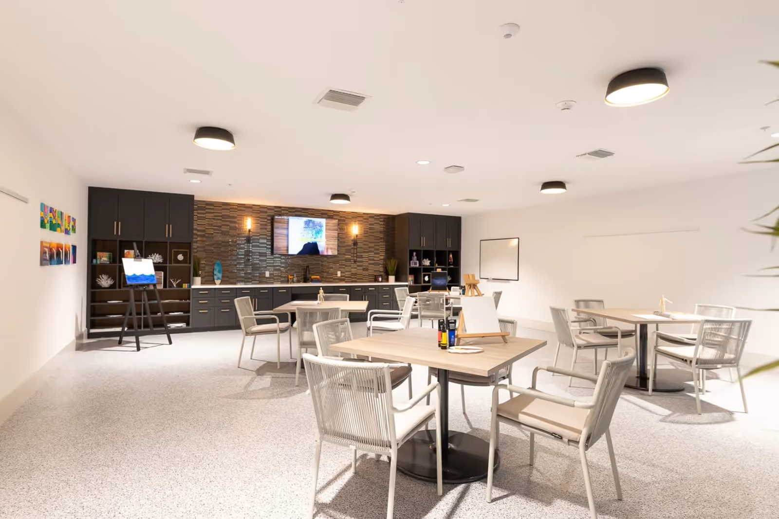 A bright, spacious activity room with several tables and chairs arranged for group activities. The room features a modern design with a tiled accent wall, dark cabinetry, a mounted TV, and art supplies including easels and paint bottles on the tables. The floor is light-colored and the ceiling has multiple round light fixtures.
