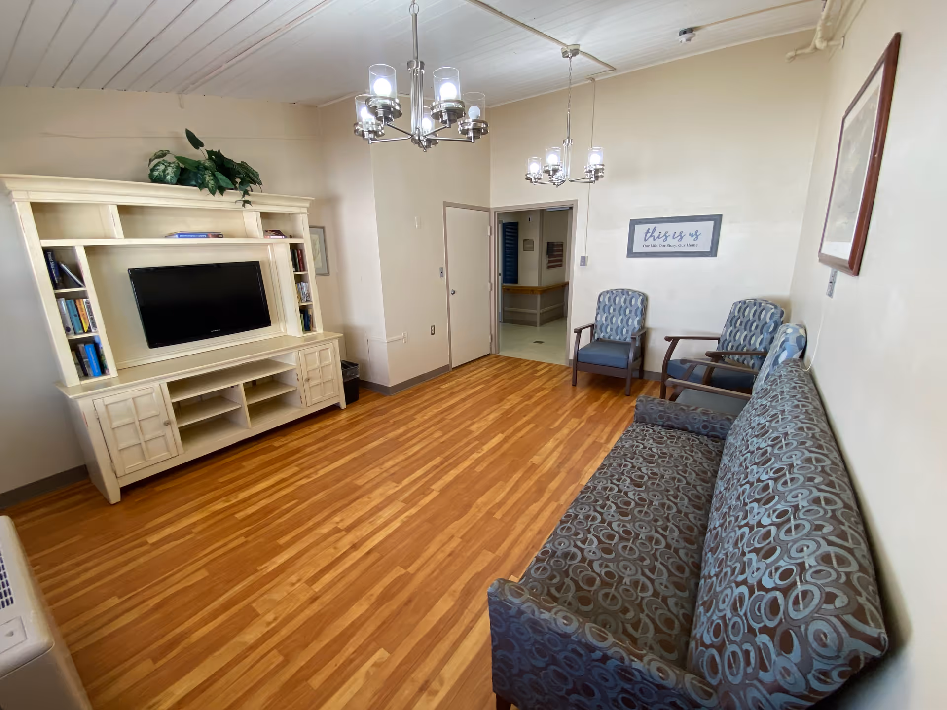Small common living room with patterned couch and chairs, a TV in a white entertainment center, wood floors, and overhead chandeliers.
