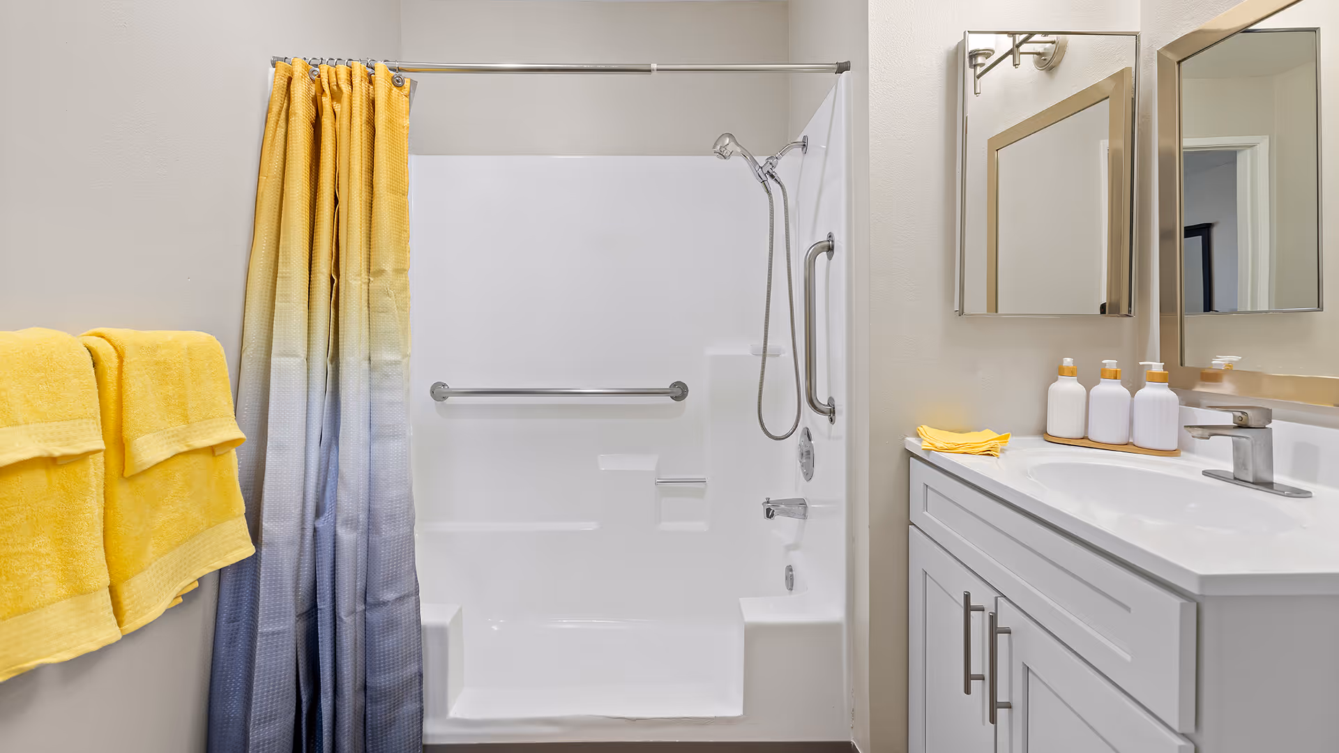 A clean bathroom featuring a white bathtub with built-in seats and grab bars, a silver showerhead, a yellow and gray ombre shower curtain, two yellow towels hanging on a wall rack, a white vanity with a sink, three white soap dispensers, and a mirror above the sink.