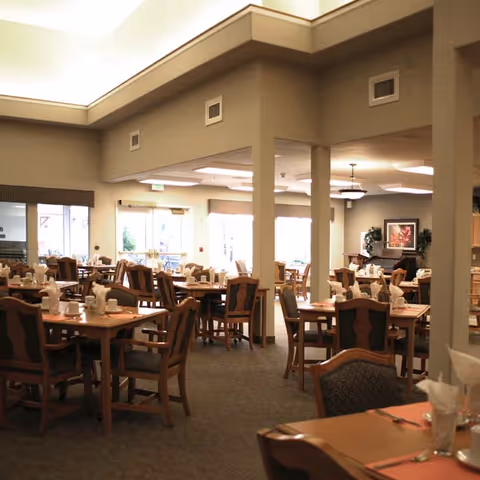 A spacious dining room with multiple wooden tables and chairs arranged neatly. Each table is set with white napkins, cups, and utensils. The room has large windows allowing natural light to brighten the space, and there are decorative plants and framed artwork on the walls.