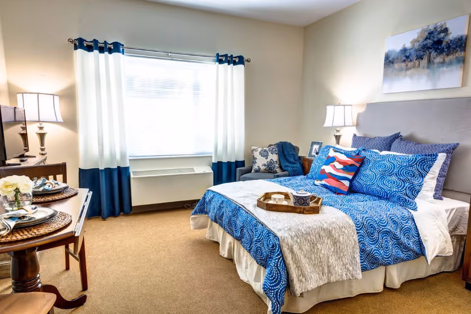 A cozy bedroom with a bed dressed in blue and white patterned bedding, a wooden side table with a lamp, a small armchair with a cushion and throw, a window with white and blue curtains, and a small dining table set for two with plates and napkins.