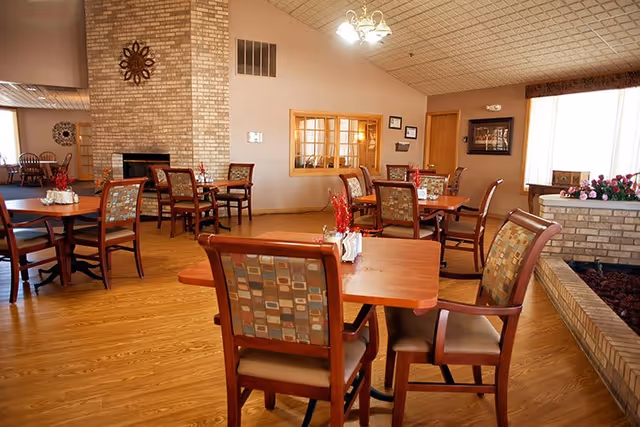 Spacious dining room with wooden tables, patterned upholstered chairs, a brick fireplace, and large windows letting in light.