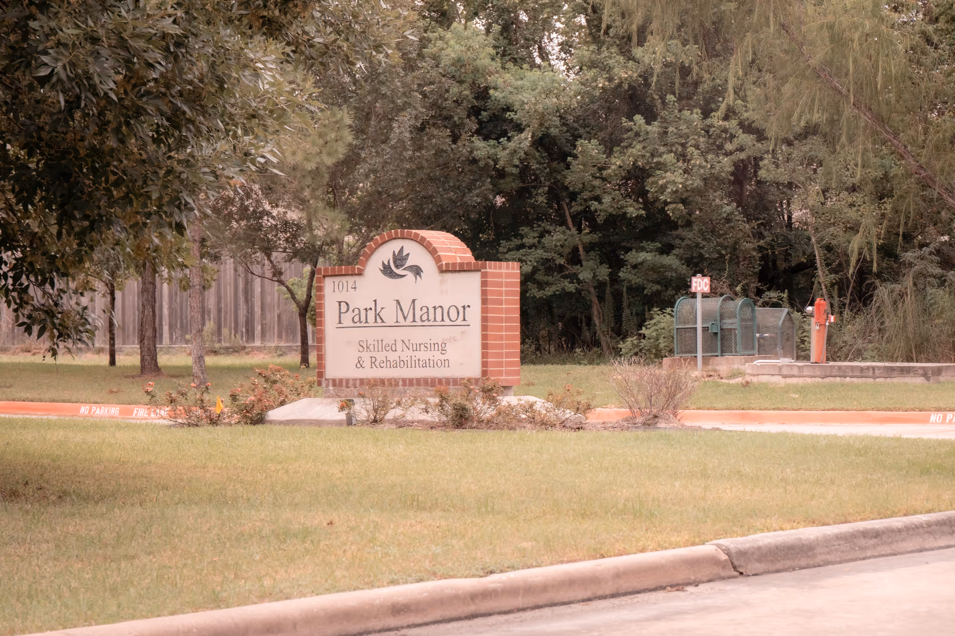 A brick and stone sign for Park Manor Skilled Nursing & Rehabilitation located on a grassy area near a driveway, surrounded by trees and bushes.