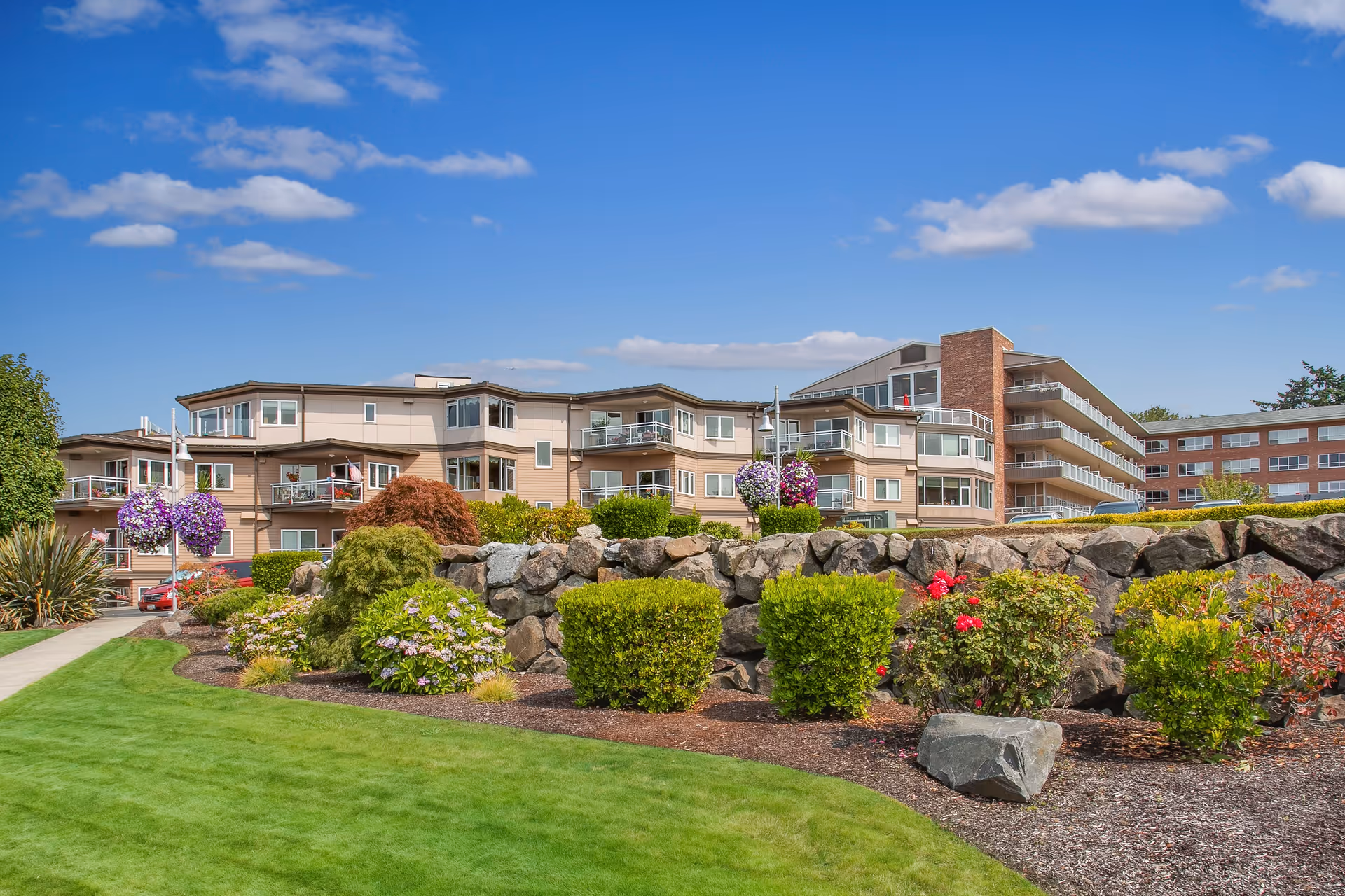 Exterior view of a multi-story retirement community building with balconies, surrounded by well-maintained landscaping including green grass, bushes, flowers, and a rock wall under a blue sky with scattered clouds.