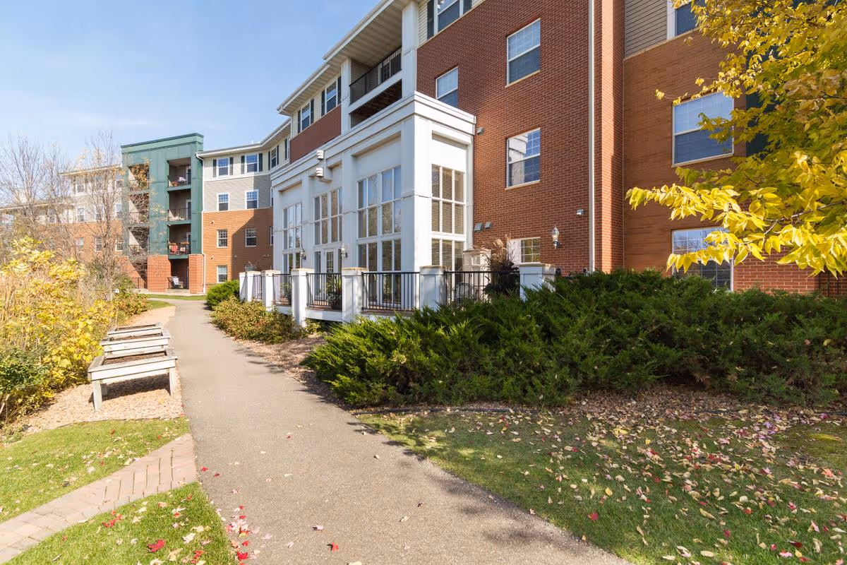 Walkway and benches leading past landscaped shrubs to the front exterior of a multi-story brick residential building.