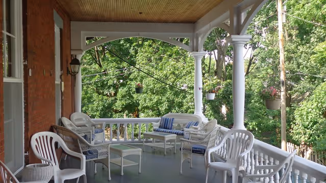 A covered porch with white wicker furniture including chairs, a loveseat with blue and white striped cushions, and small tables. The porch has white railings and columns, a wooden ceiling, and overlooks green trees in the background.