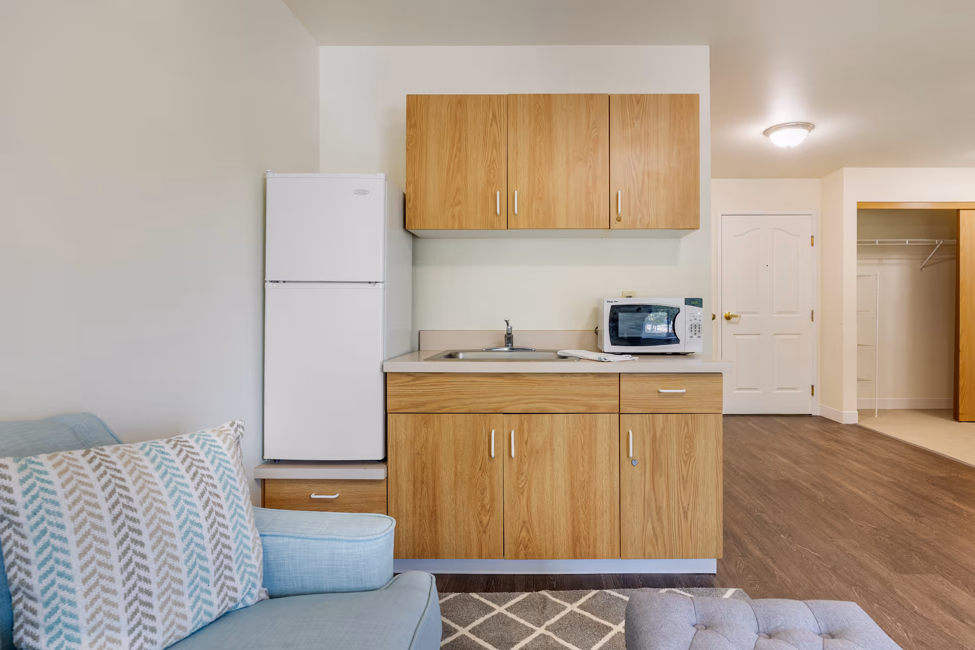 Small kitchenette area with wooden cabinets, a white refrigerator, a microwave on the countertop, and a sink. To the left is a light blue armchair with a patterned pillow. The floor is wood, and there is a white door and an open closet in the background.