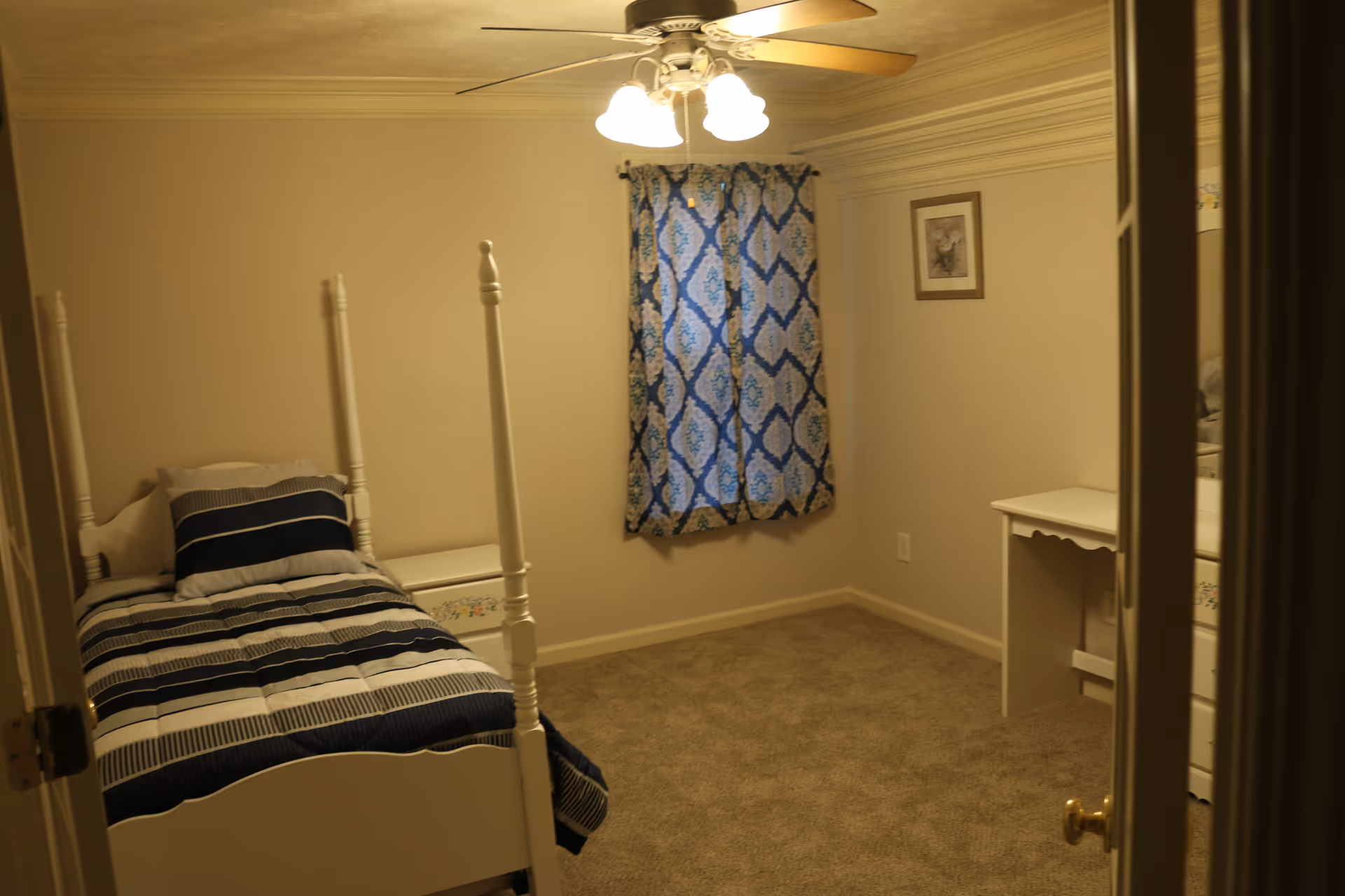 Small bedroom with a single four-poster bed with blue-striped bedding, a patterned curtain, ceiling fan lighting, and a small desk/dresser.