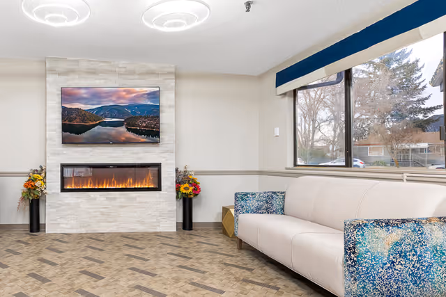 A cozy living room area with a modern electric fireplace set in a white stone wall. Above the fireplace is a large flat-screen TV displaying a scenic landscape of mountains and a lake at sunset. On either side of the fireplace are tall black vases with colorful flower arrangements. To the right is a beige sofa with blue patterned armrests, positioned near a large window with a blue valance, showing trees and houses outside.