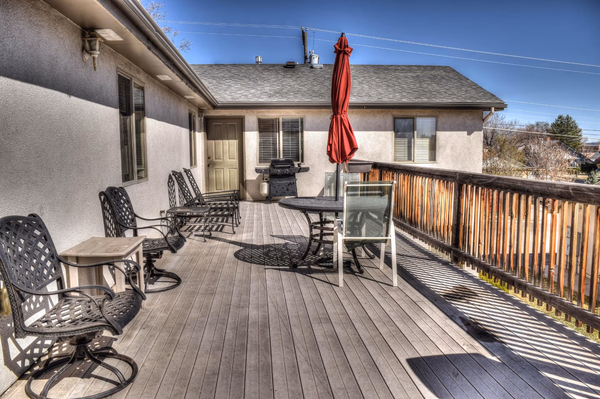 Outdoor wooden deck area with several black metal chairs lined up against the building wall, a round table with a closed red umbrella, a grill, and a wooden railing overlooking a residential neighborhood under a clear blue sky.