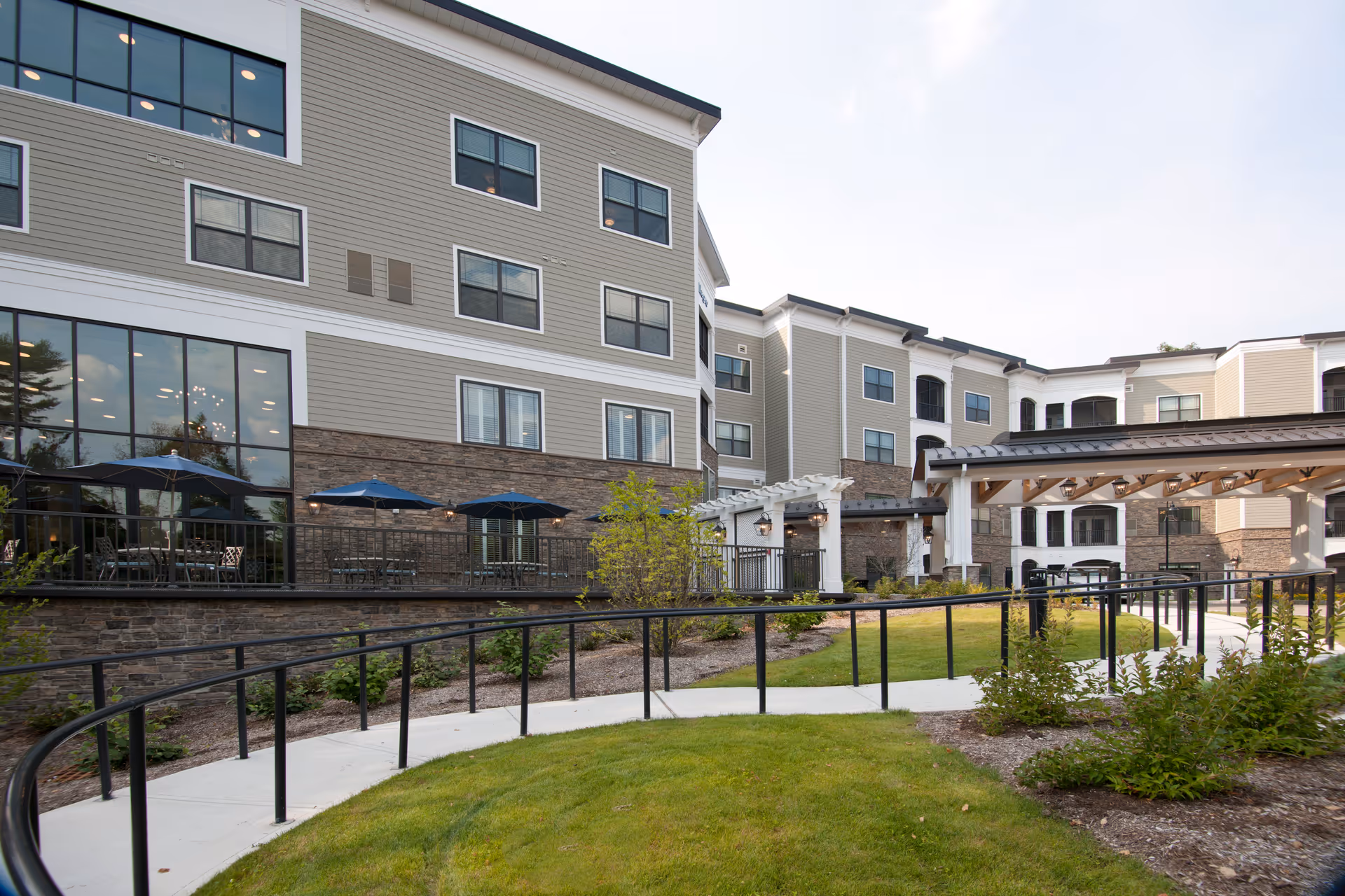 Exterior view of a senior living facility building with multiple floors, large windows, and a patio area with tables and umbrellas. There is a paved walkway with black railings surrounded by green grass and landscaped plants.