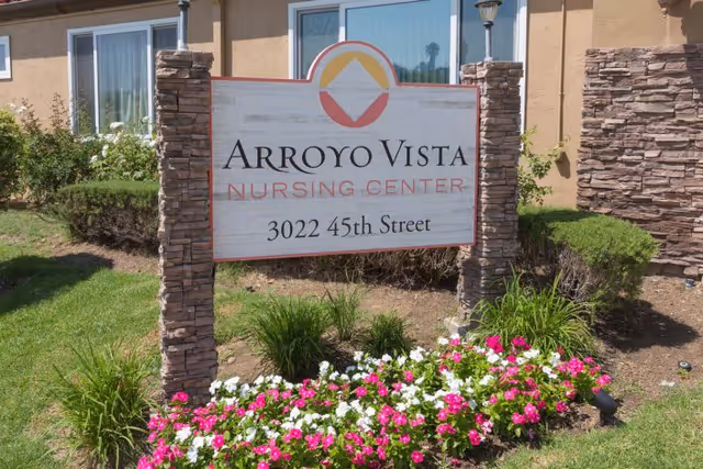 Outdoor view of a sign for Arroyo Vista Nursing Center at 3022 45th Street, surrounded by green bushes and colorful flowers in front of a beige building with stone accents.
