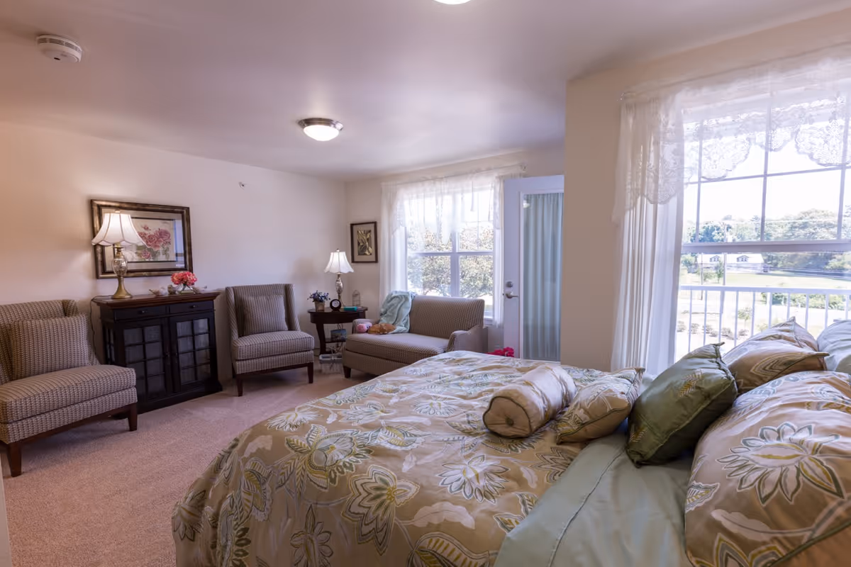 Sunlit bedroom with a floral-patterned bed, seating area with upholstered chairs and a cabinet, and large windows overlooking the outdoors.