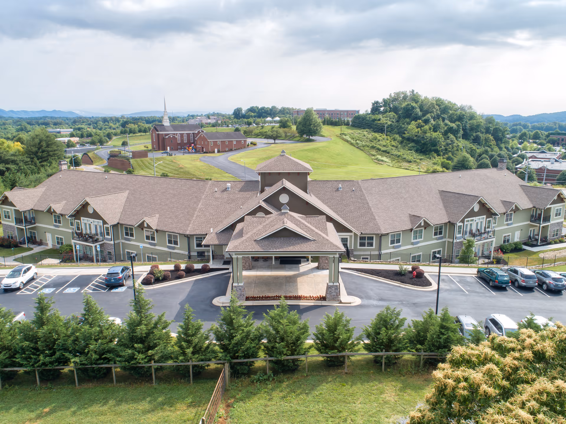 Aerial view of Dominion Senior Living facility showing a large, single-story building with a covered entrance and multiple balconies. The building is surrounded by a parking lot with several cars parked. In the background, there is a green hill, a church with a tall steeple, and a scenic landscape with trees and distant mountains under a partly cloudy sky.