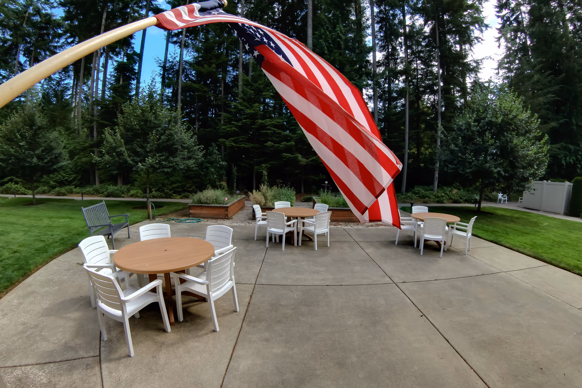 Outdoor patio area with three round tables each surrounded by white plastic chairs. An American flag is prominently displayed in the foreground, waving on a curved flagpole. The patio is surrounded by green grass, trees, and some raised garden beds in the background.