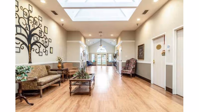 A spacious hallway in a healthcare center with wooden flooring, beige walls, and a high ceiling featuring skylights. The hallway is furnished with cushioned chairs and sofas along the walls, a wooden coffee table with plants in the center, and decorative wall art including a tree with the word 'Respect'. The far end has large glass doors allowing natural light to enter.