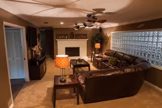 Cozy living room with brown leather sectional sofa, wooden coffee table, side tables with lamps, ceiling fans, and a fireplace. The room has beige walls and carpet, with a glass block window allowing natural light.