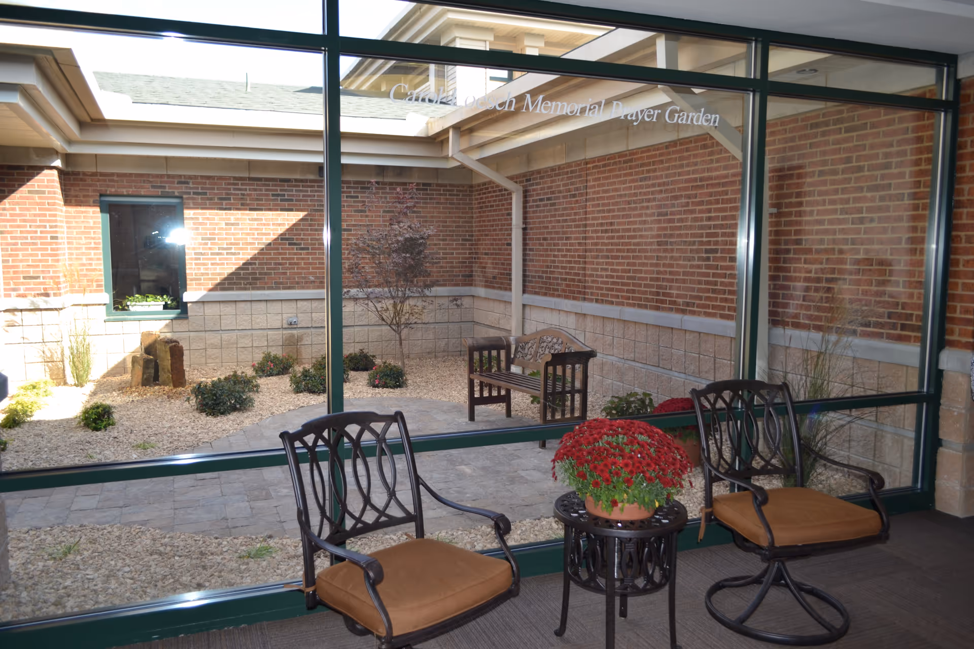 Indoor seating area with two cushioned chairs and a small round table holding a potted red flower, looking out through large glass windows onto a small enclosed garden with a bench, plants, and a brick wall.