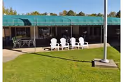 Outdoor lawn and patio area in front of a single-story building with white Adirondack chairs under a green awning.